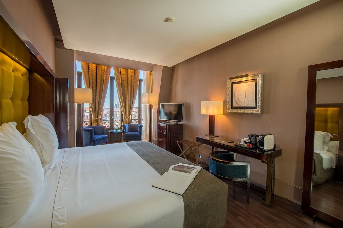 A hotel room with a large bed, a desk with a television, a chair, and a desk lamp.