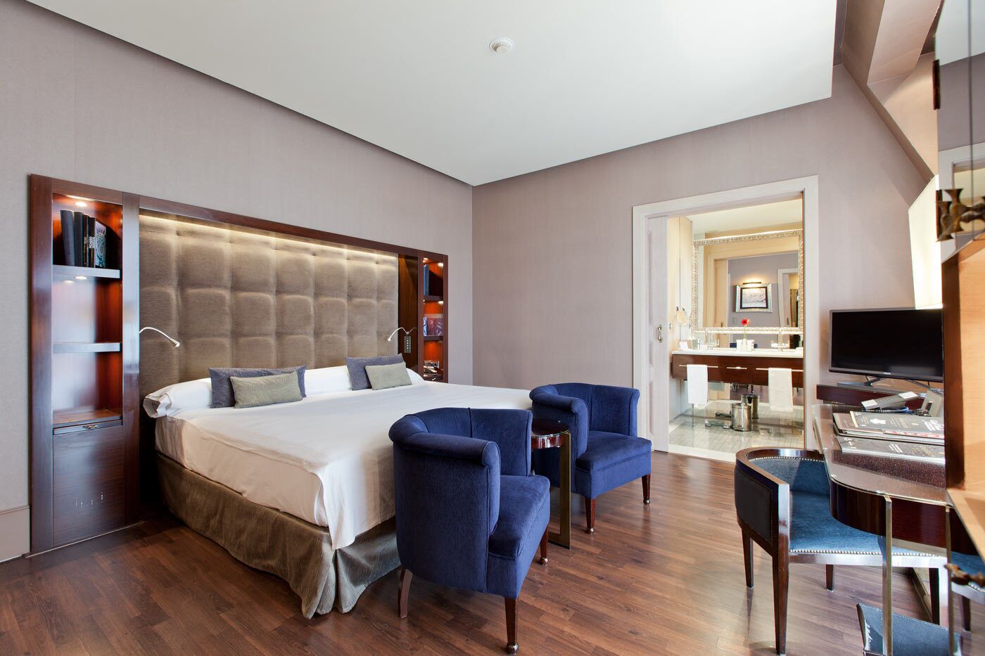 Een hotelkamer met een groot bed, twee blauwe fauteuils, een bureau met een stoel, een televisie en een zichtbare badkamer door een open deur.