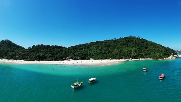 A tropical island with a white sandy beach, clear turquoise waters, and several boats. - Morro das Pedras Clube Hotel (Florianópolis)