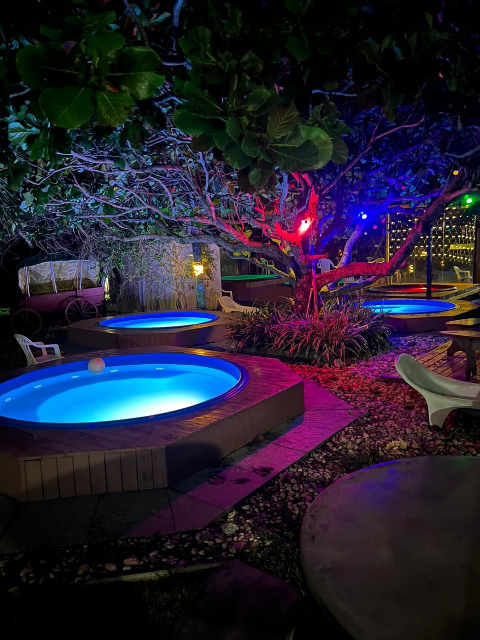 Escena nocturna al aire libre con múltiples jacuzzis iluminados por luces de colores, rodeados de follaje y un camino de piedra.