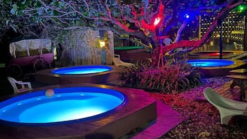 Escena nocturna al aire libre con múltiples jacuzzis iluminados por luces de colores, rodeados de follaje y un camino de piedra.
