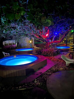 A nighttime outdoor setting with multiple hot tubs illuminated by colored lights, surrounded by foliage and a stone pathway. - Morro das Pedras Clube Hotel (Florianópolis)