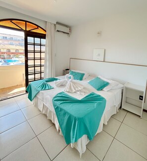 A hotel room with a bed, a nightstand, a window with a view of a pool and buildings, and an air conditioning unit. - Morro das Pedras Clube Hotel (Florianópolis)