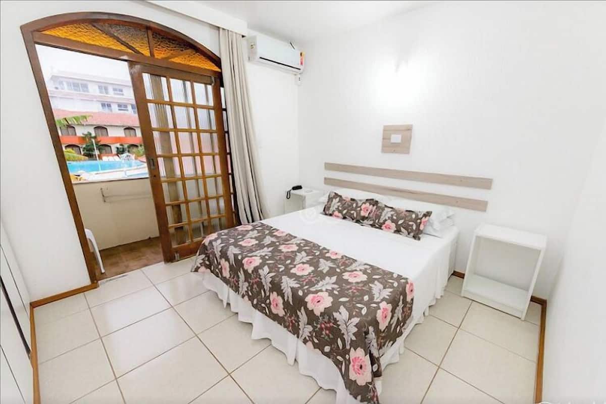 A bedroom with a bed, a bedside table, a nightstand, a window with a view of a building and a pool.