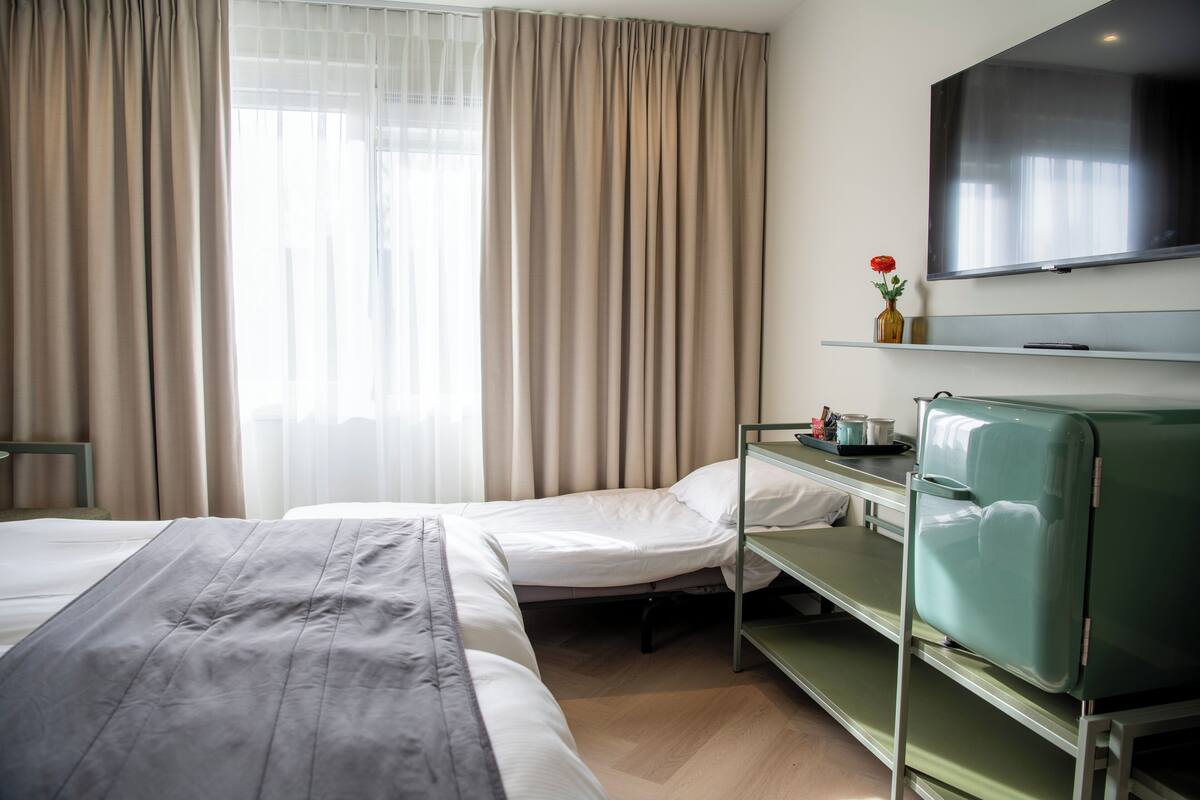 A hotel room with two beds, a television, a bedside table with a vase and a tray, and a window with curtains.