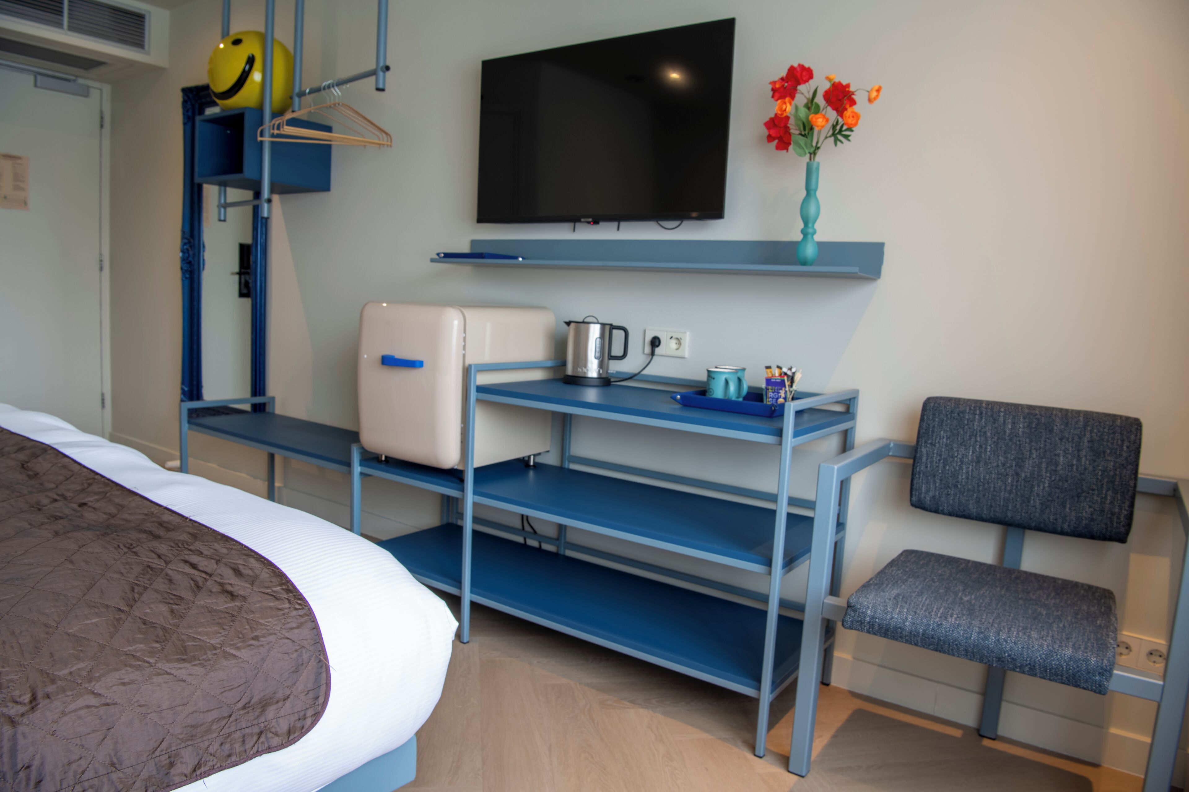 A hotel room with a bed, a desk with a kettle, a chair, a TV, and a vase with flowers.