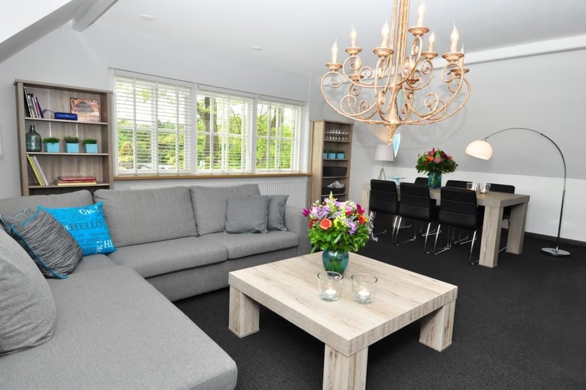 A modern living room with a grey sofa, a wooden coffee table, a chandelier, and a dining area with black chairs.