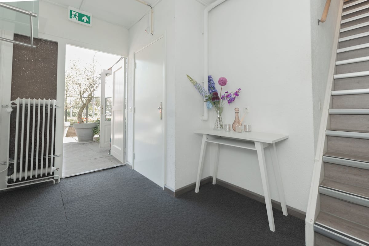 A hallway with a radiator, a white table with a vase of flowers, and a staircase.