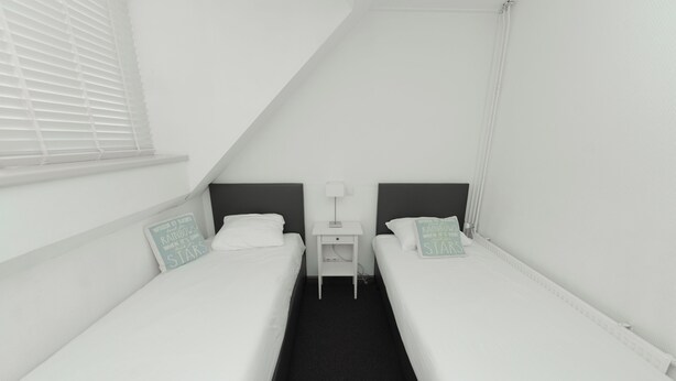 A twin room with two single beds, a small bedside table with a lamp, and a window with blinds. - Boutique Hotel Herbergh Amsterdam Airport (Badhoevedorp)