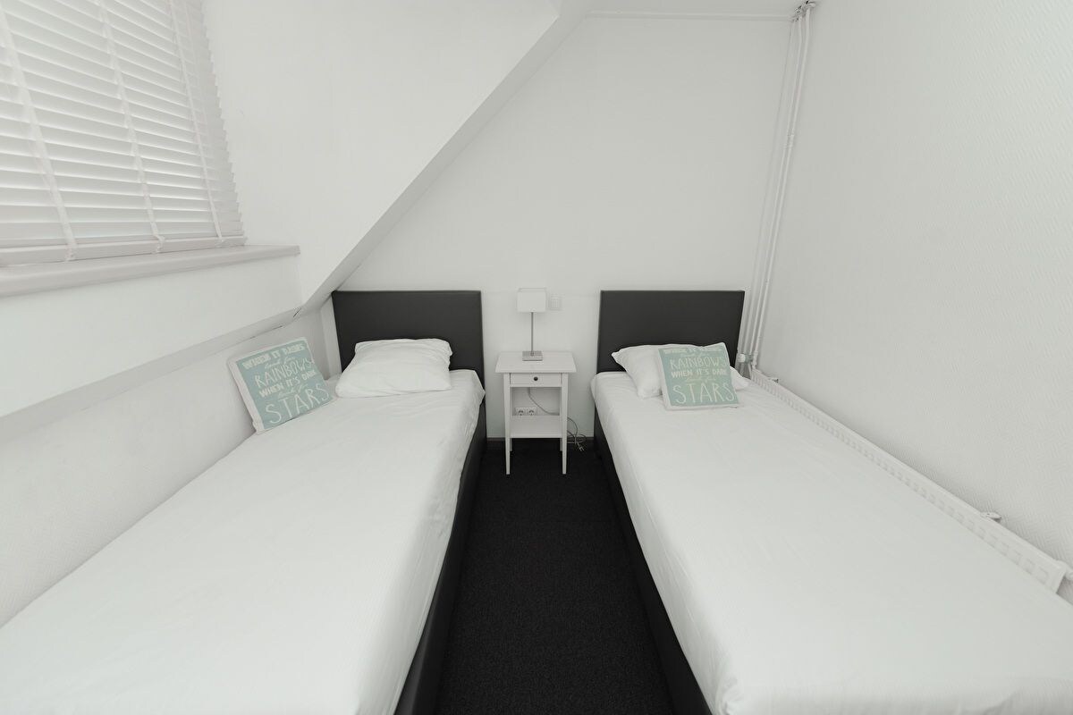 A twin room with two single beds, a small bedside table with a lamp, and a window with blinds.