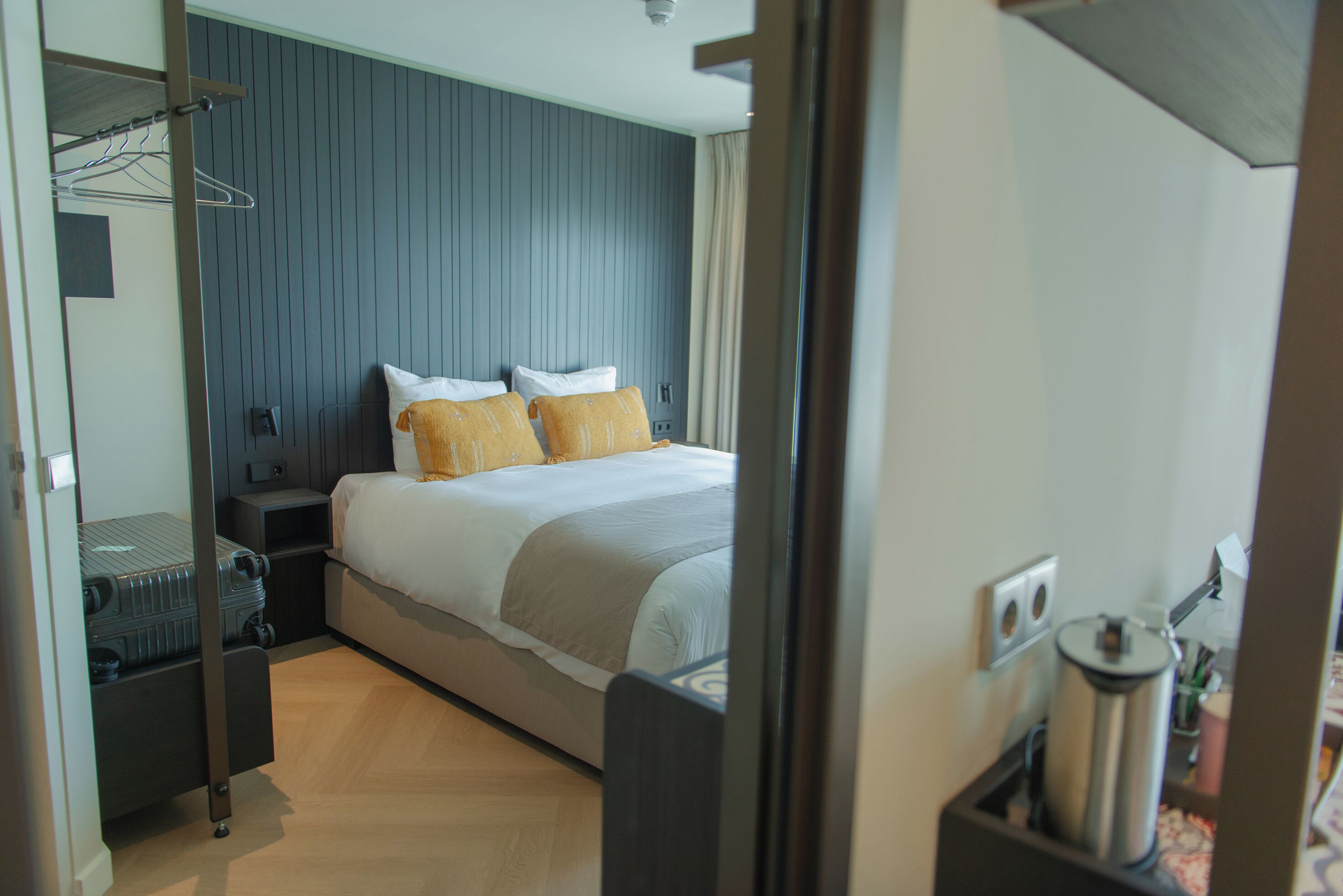 A hotel room with a bed, a nightstand, a suitcase, and a wall-mounted heater.