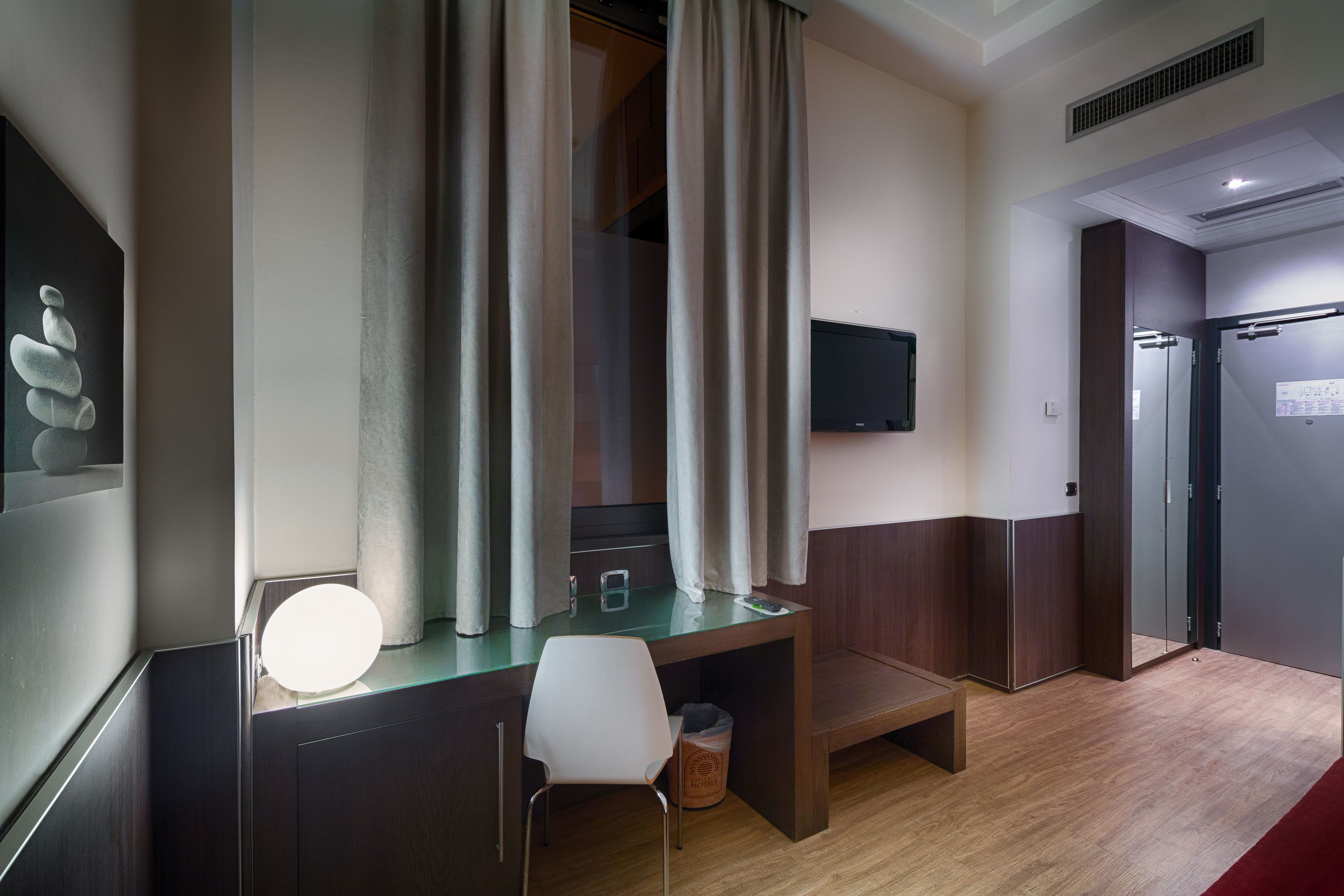 A modern hotel room with a wooden desk, a white chair, a flat-screen TV mounted on the wall, and a red carpet.