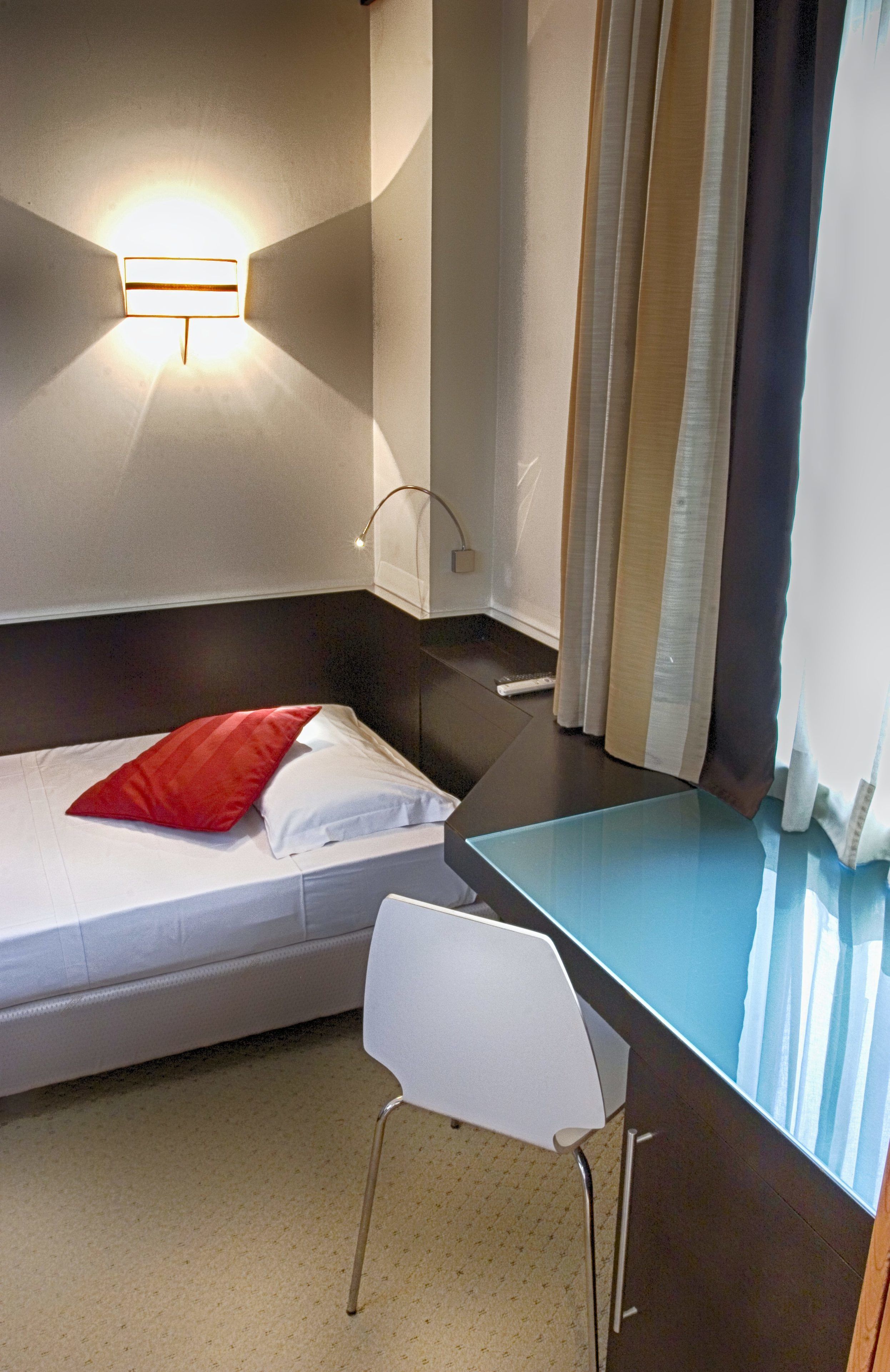 A small, neatly arranged bedroom with a single bed, a bedside table, a chair, and a wall-mounted light fixture.