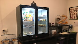 Una kitchenette con un refrigerador de doble puerta repleto de bebidas y alimentos, una cafetera y un frutero en la encimera.