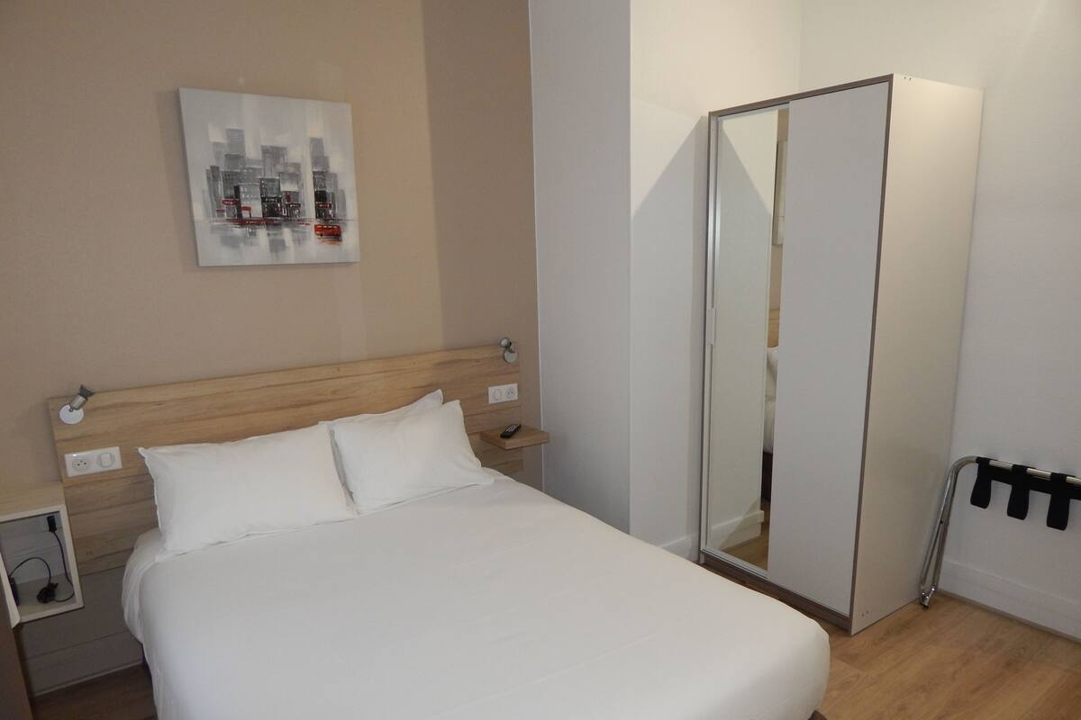 A hotel room with a bed, a nightstand, a mirror, and a cityscape artwork on the wall.