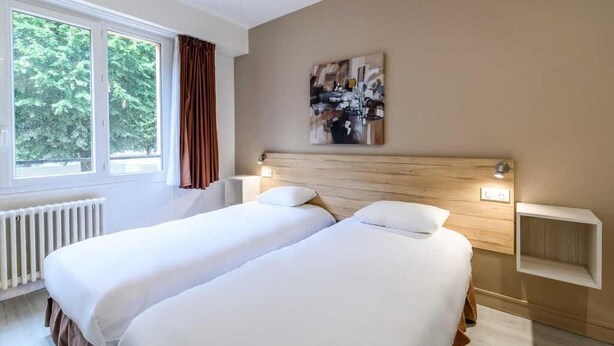 Habitación de hotel con dos camas, una ventana con cortinas, un radiador, un cuadro en la pared y una mesita de noche con lámpara.