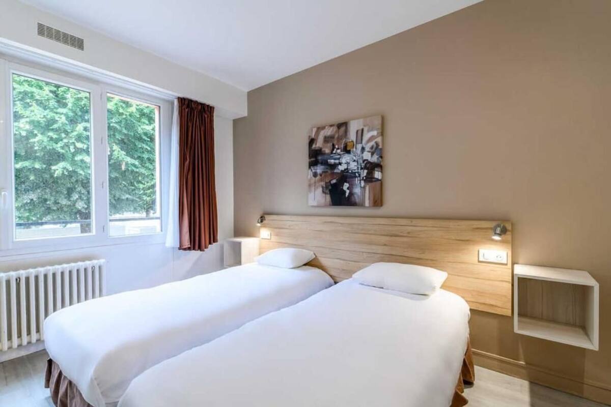 A hotel room with two beds, a window with curtains, a radiator, a painting on the wall, and a bedside table with a lamp.