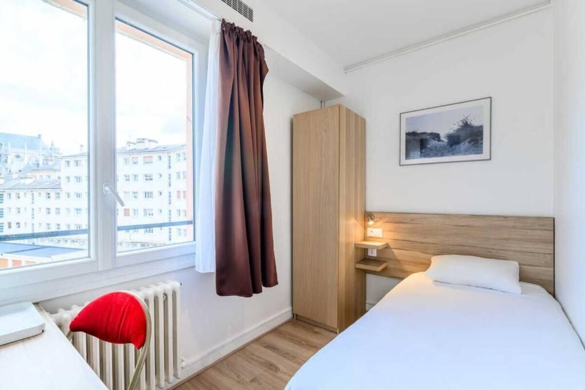 A hotel room with a bed, a nightstand, a red chair, a radiator, a window with curtains, a wardrobe, and a framed picture on the wall.