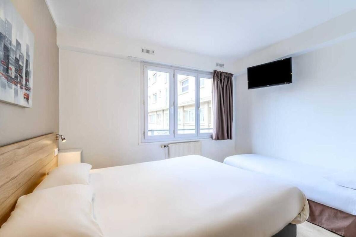A hotel room with a bed, a window with curtains, a wall-mounted TV, and a radiator.