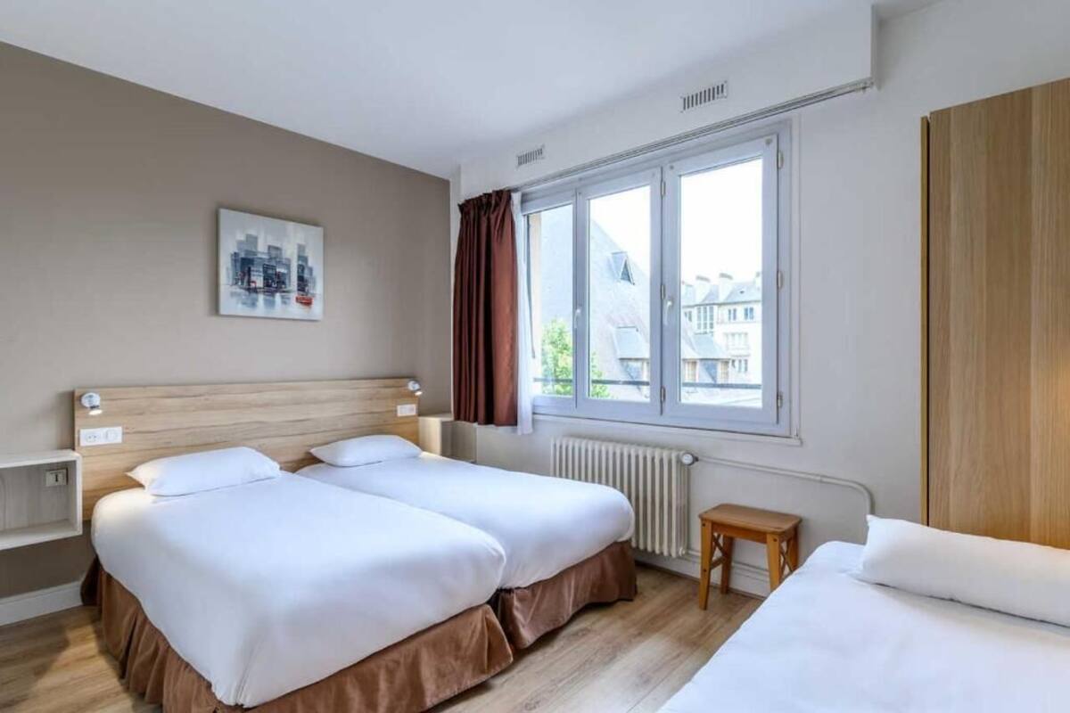 A hotel room with two beds, a window with curtains, a radiator, and a small table.