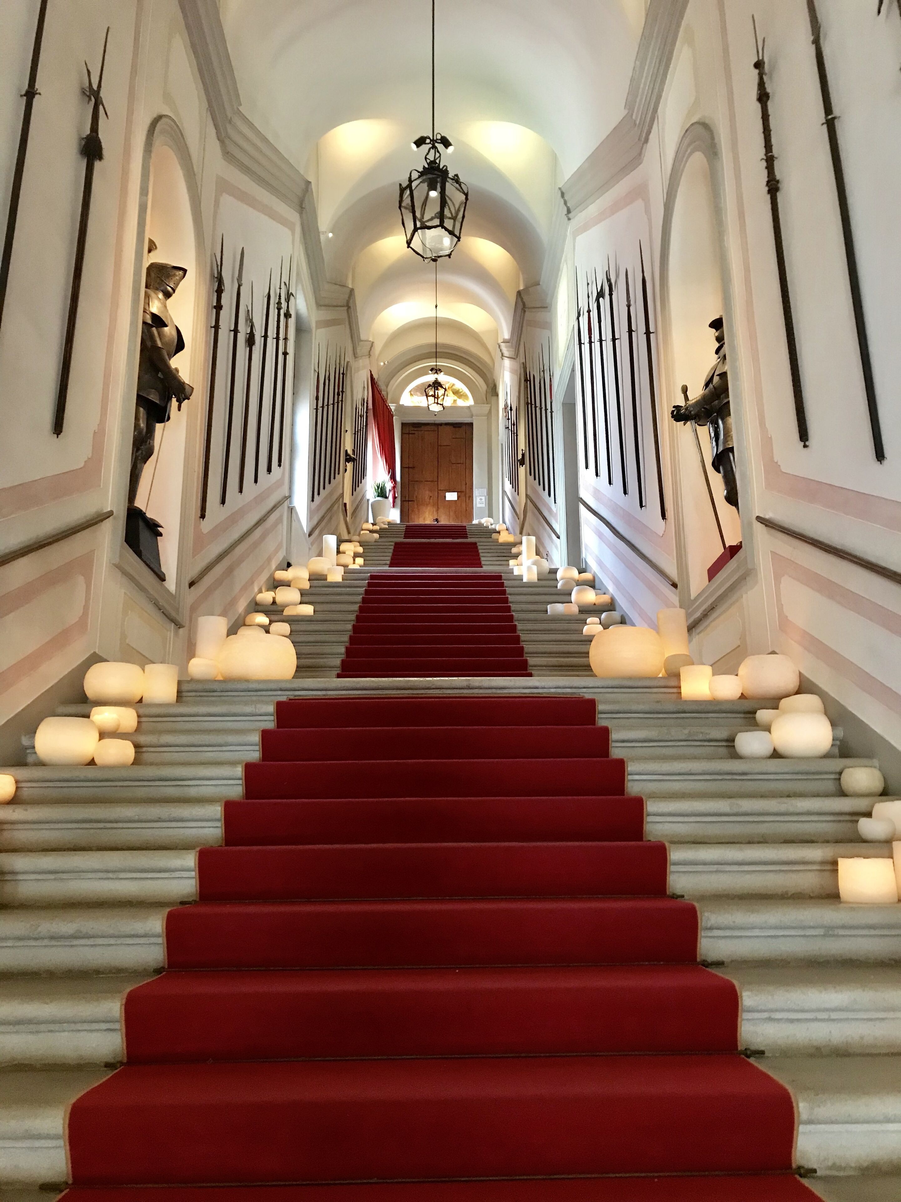 A grand staircase with red carpet, flanked by wall-mounted weapons and a chandelier.