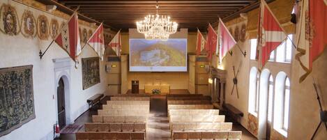 Un salón de conferencias con bancos y mesas de madera, una araña, banderas y una pantalla de proyección que muestra un castillo.
