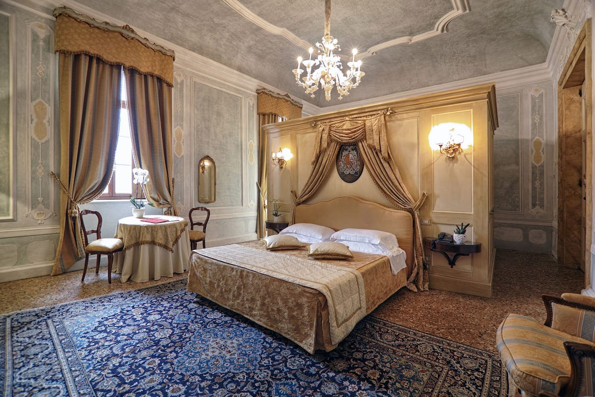 A luxurious bedroom with a large bed, ornate curtains, a chandelier, and a patterned rug.