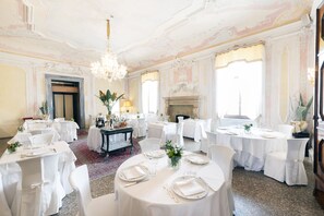 A formal dining room with round tables, white chairs, chandeliers, and a fireplace. - Castelbrando (Cison di Valmarino)