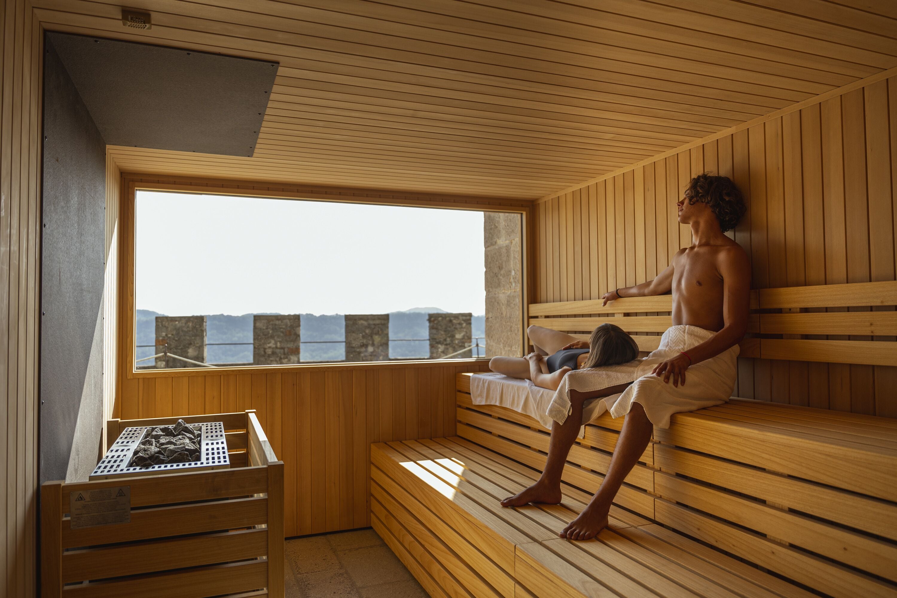 A sauna room with wooden benches, a large window, and a stone container.