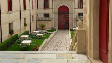 Un patio con una puerta roja central, rodeado de mesas y sillas, y un edificio con contraventanas rojas y ventanas de arco.
