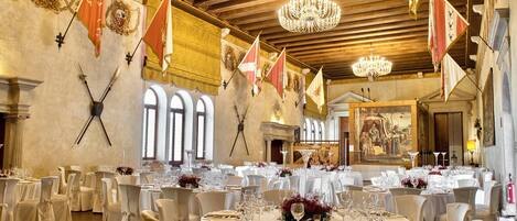 Un salón de banquetes con mesas redondas dispuestas para cenar, decorado con manteles blancos y centros de mesa florales rojos.