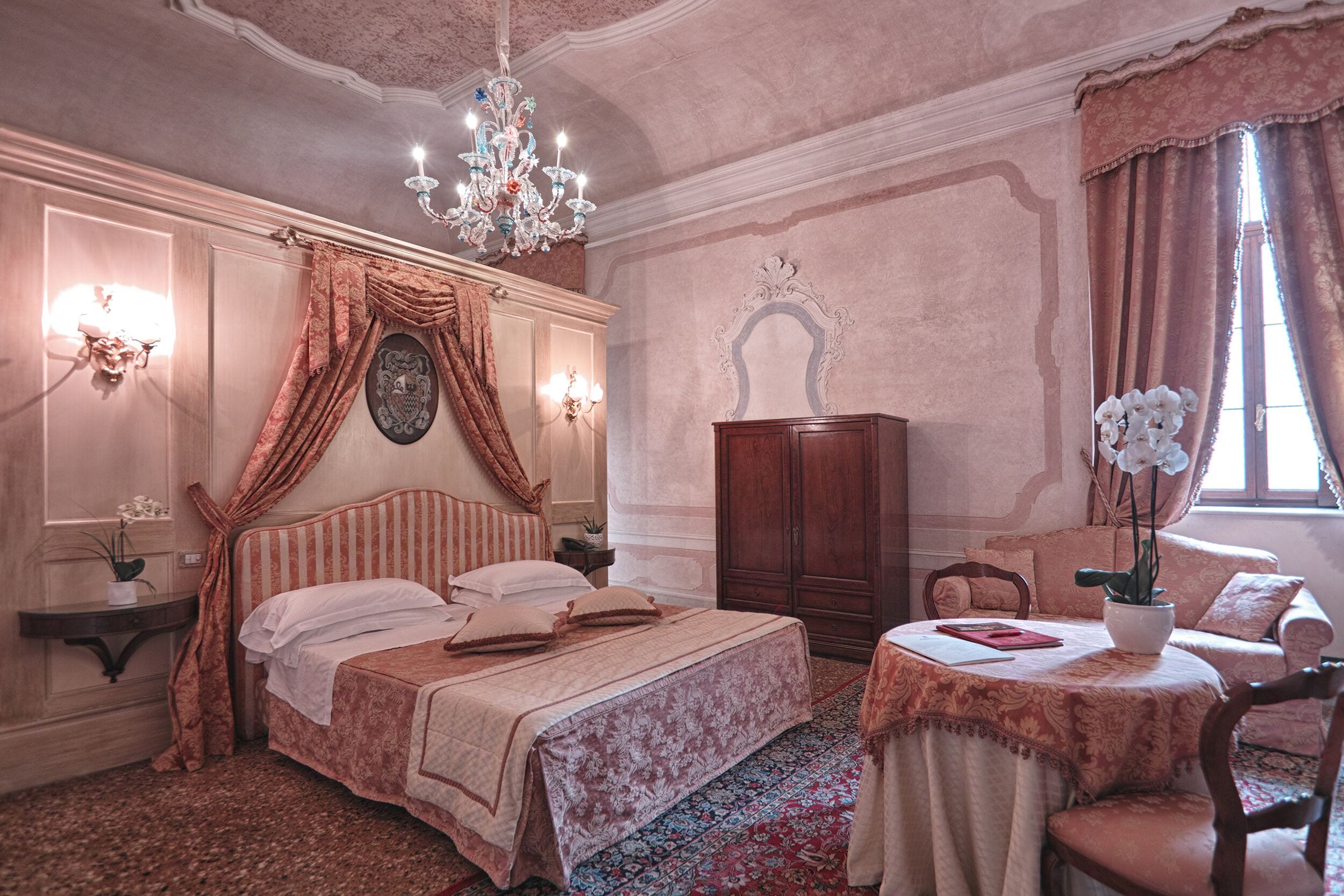 A bedroom with a large bed, a chandelier, a wooden wardrobe, a small table with a vase, and a window with curtains.