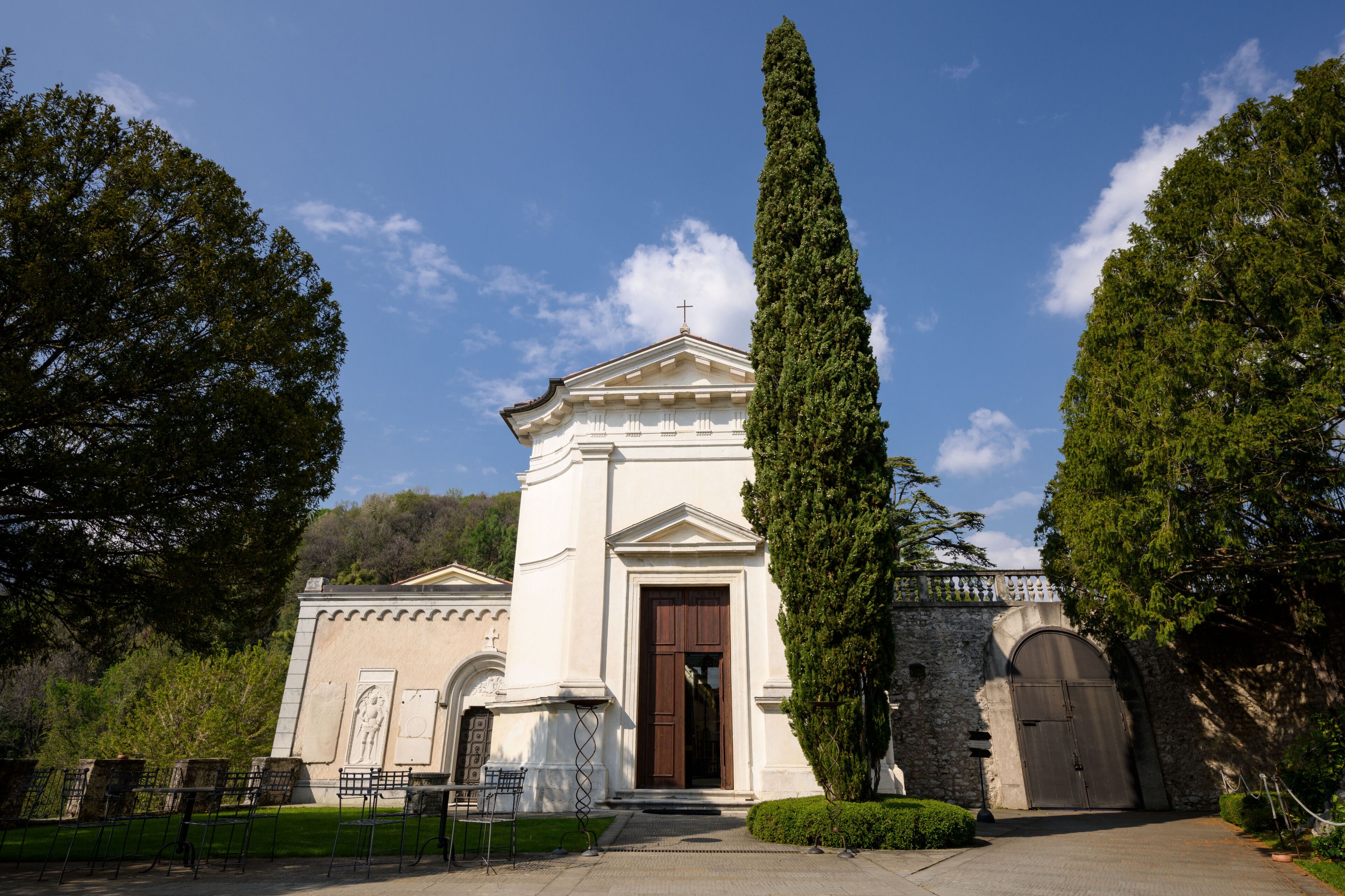 Un edificio bianco con un ingresso centrale, affiancato da due alberi alti e un muro di pietra.