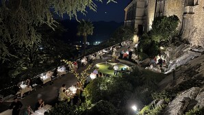 An outdoor dining area with tables and chairs, surrounded by trees and a stone building.