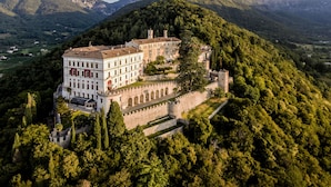 A historic building with multiple wings, surrounded by lush greenery and mountains in the background.