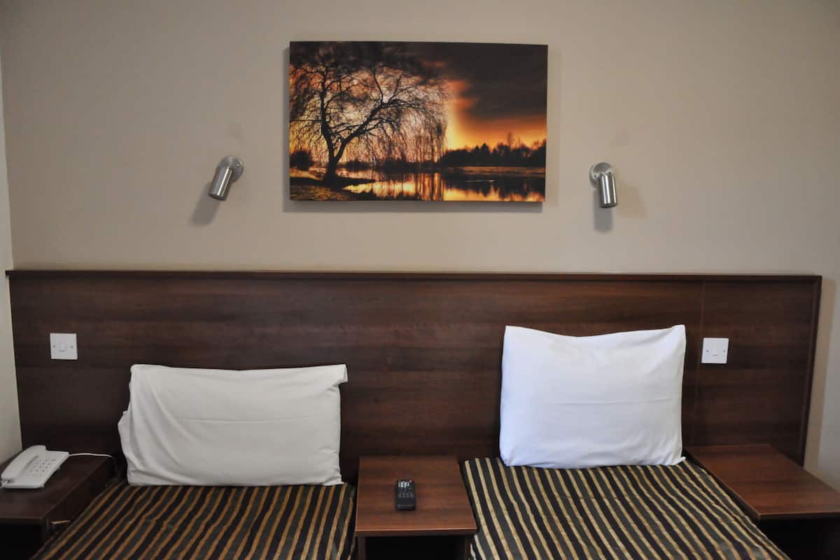 Hotelkamer met een houten hoofdeinde, twee bedden met gestreept beddengoed, een schilderij van een zonsondergang boven een meer en een nachtkastje met een telefoon en afstandsbediening.