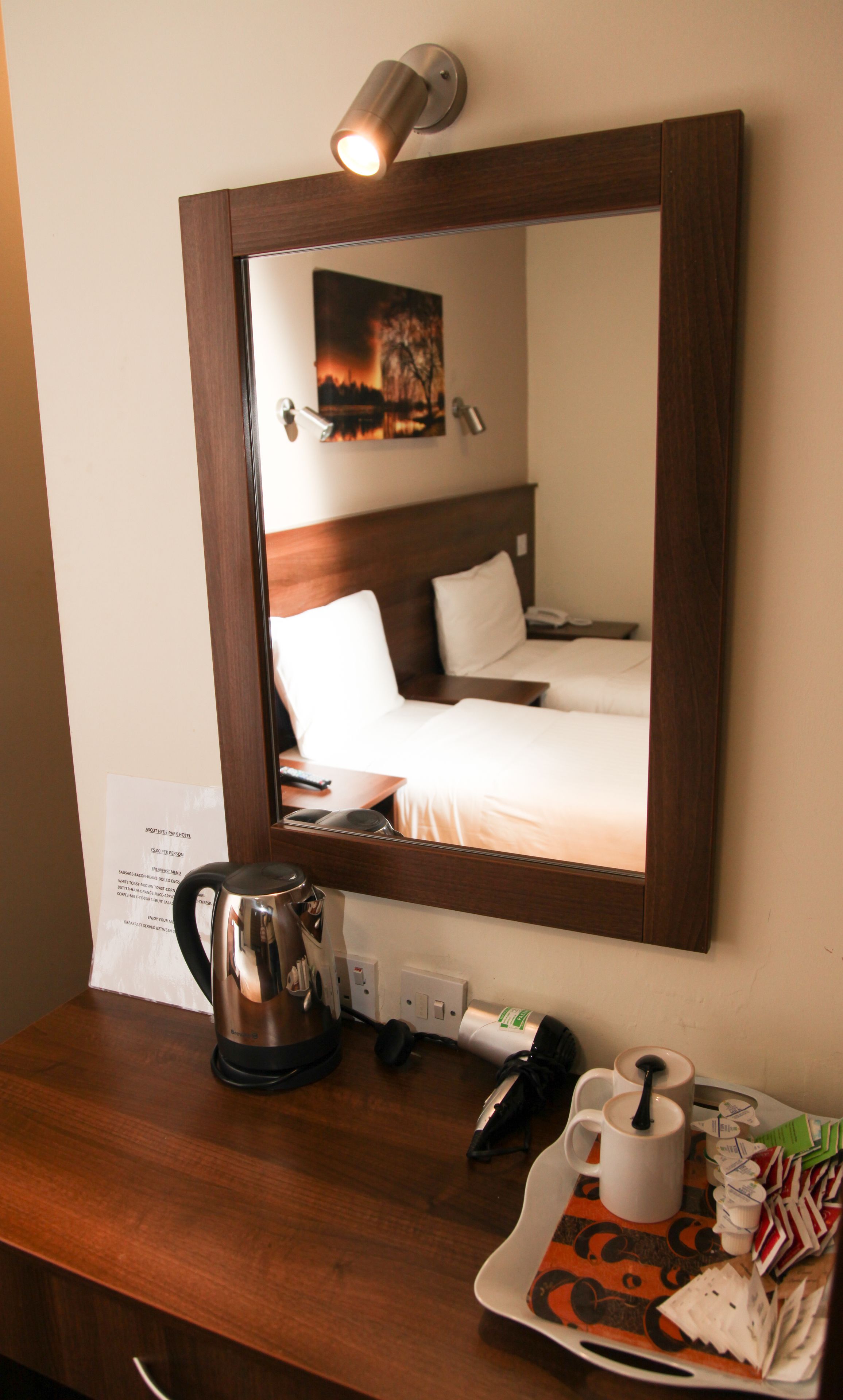 A hotel room with a wooden headboard, a bed with white linens, a bedside table with a kettle, a cup, and a tray with breakfast items.
