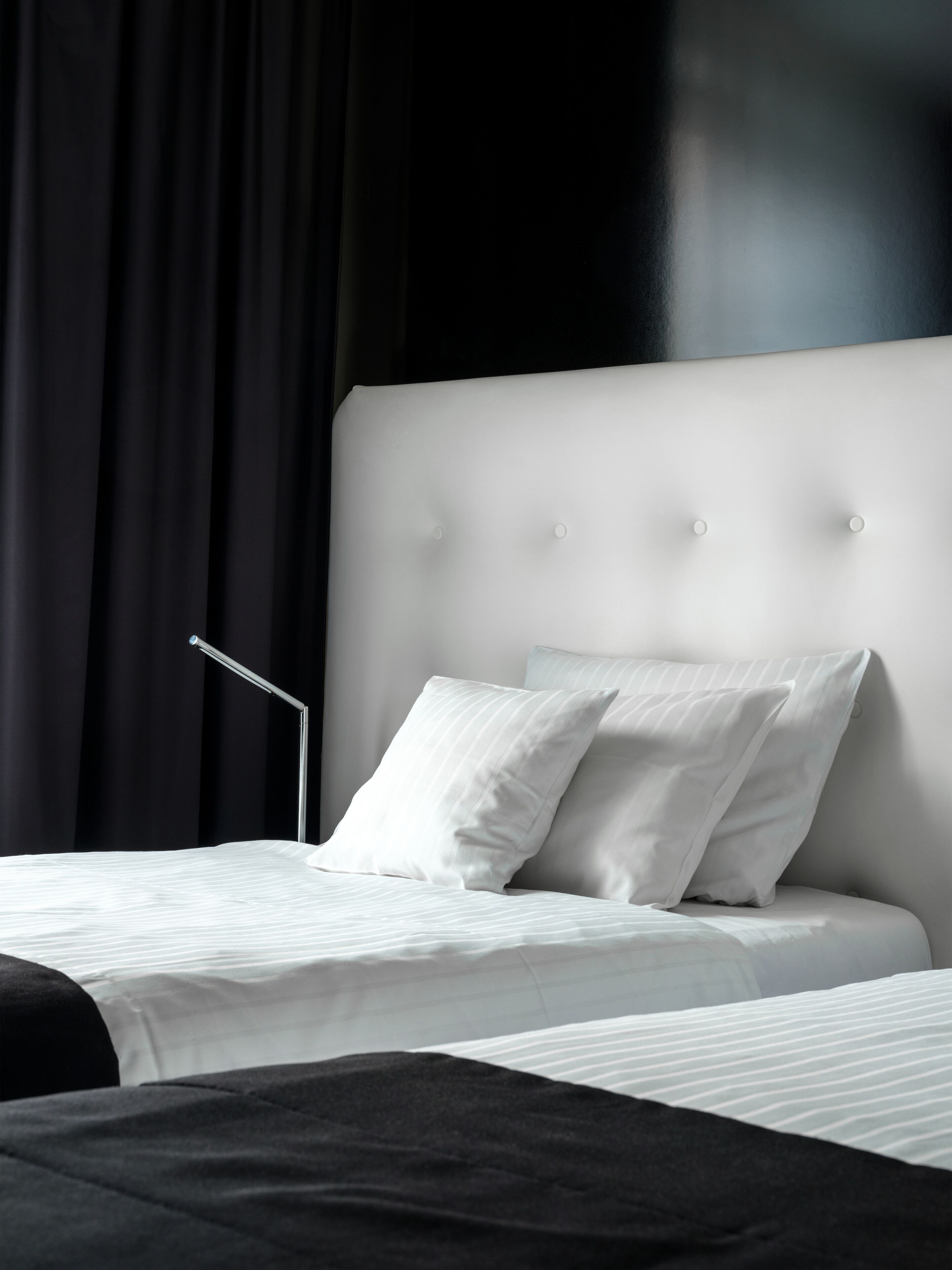 A modern bedroom with a white headboard, two pillows, a bedside lamp, and a black bedspread.