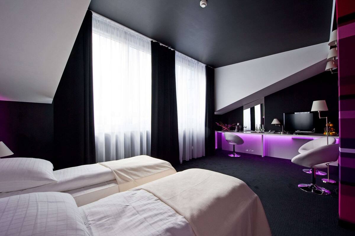 Una habitación de hotel moderna con dos camas, un escritorio con televisor y una zona de bar iluminada en morado.