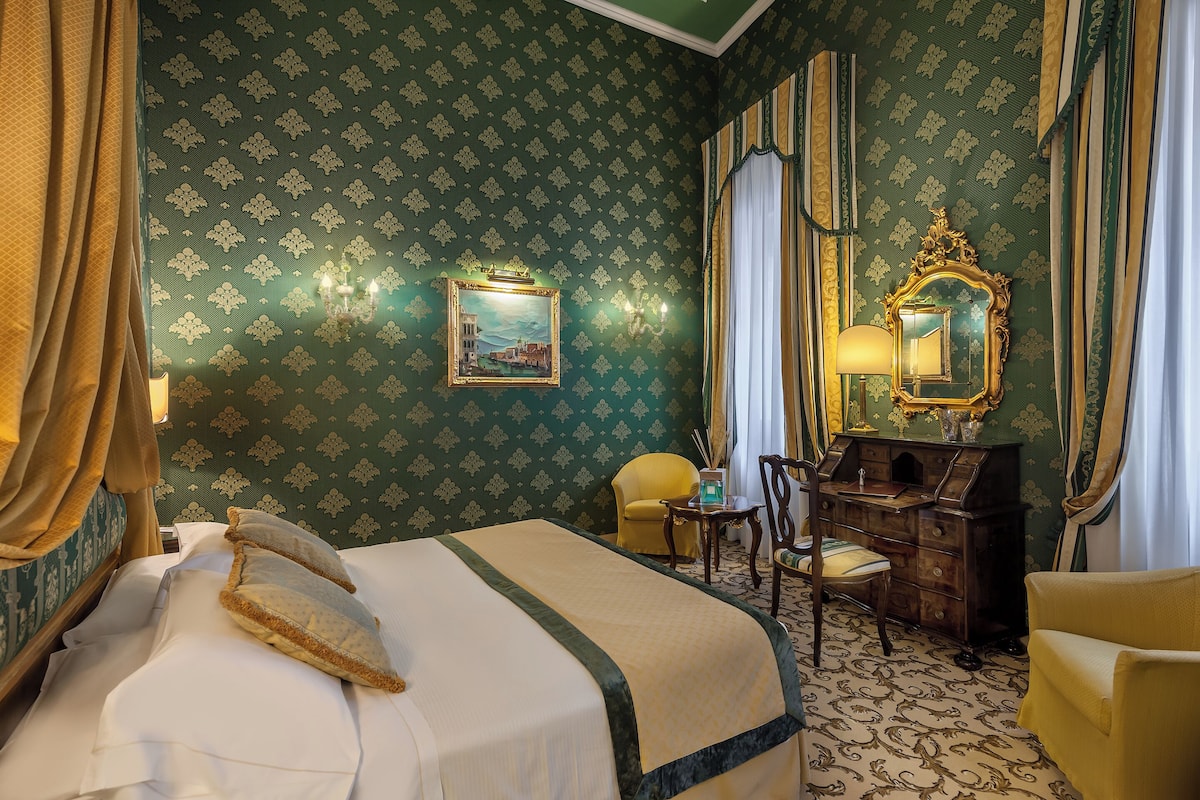 Una habitación de hotel con cama, escritorio, silla, espejo y un cuadro en la pared.