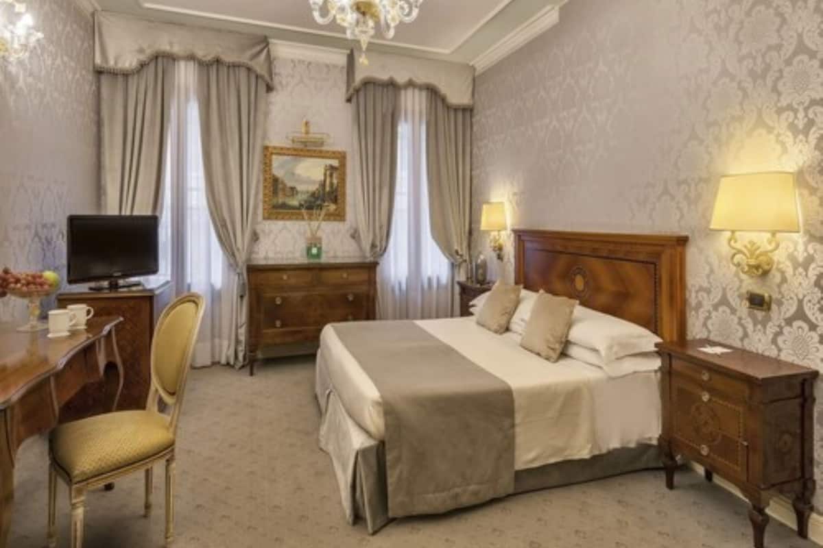 Habitación de hotel con cama, escritorio con computadora, silla, televisor y candelabro.