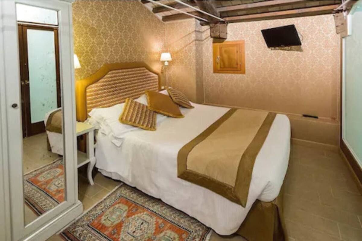 Una habitación de hotel con cama, mesita de noche, lámpara, televisor y una alfombra estampada.