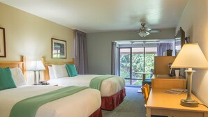 Habitación de hotel con dos camas, un escritorio, una silla, una lámpara, ventilador de techo y un ventanal con vistas a la vegetación.