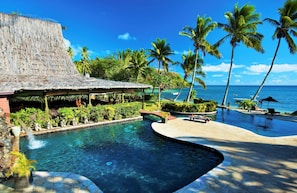 A pool area with a thatched-roof structure, palm trees, and a view of the ocean. - Beqa Lagoon Resort (Beqa Island)