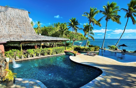 A pool area with a thatched-roof structure, palm trees, and a view of the ocean.