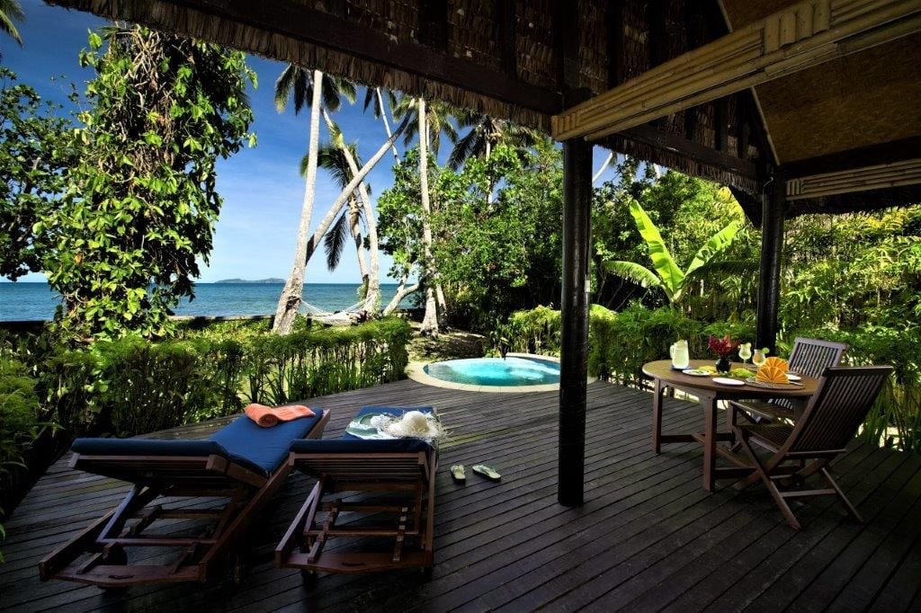 A wooden deck with lounge chairs and a table, overlooking a pool and tropical vegetation.