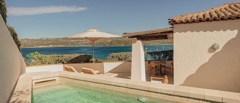 Una piscina cristalina con vistas al mar, una pérgola con toldo y un patio con sillas y una mesa.