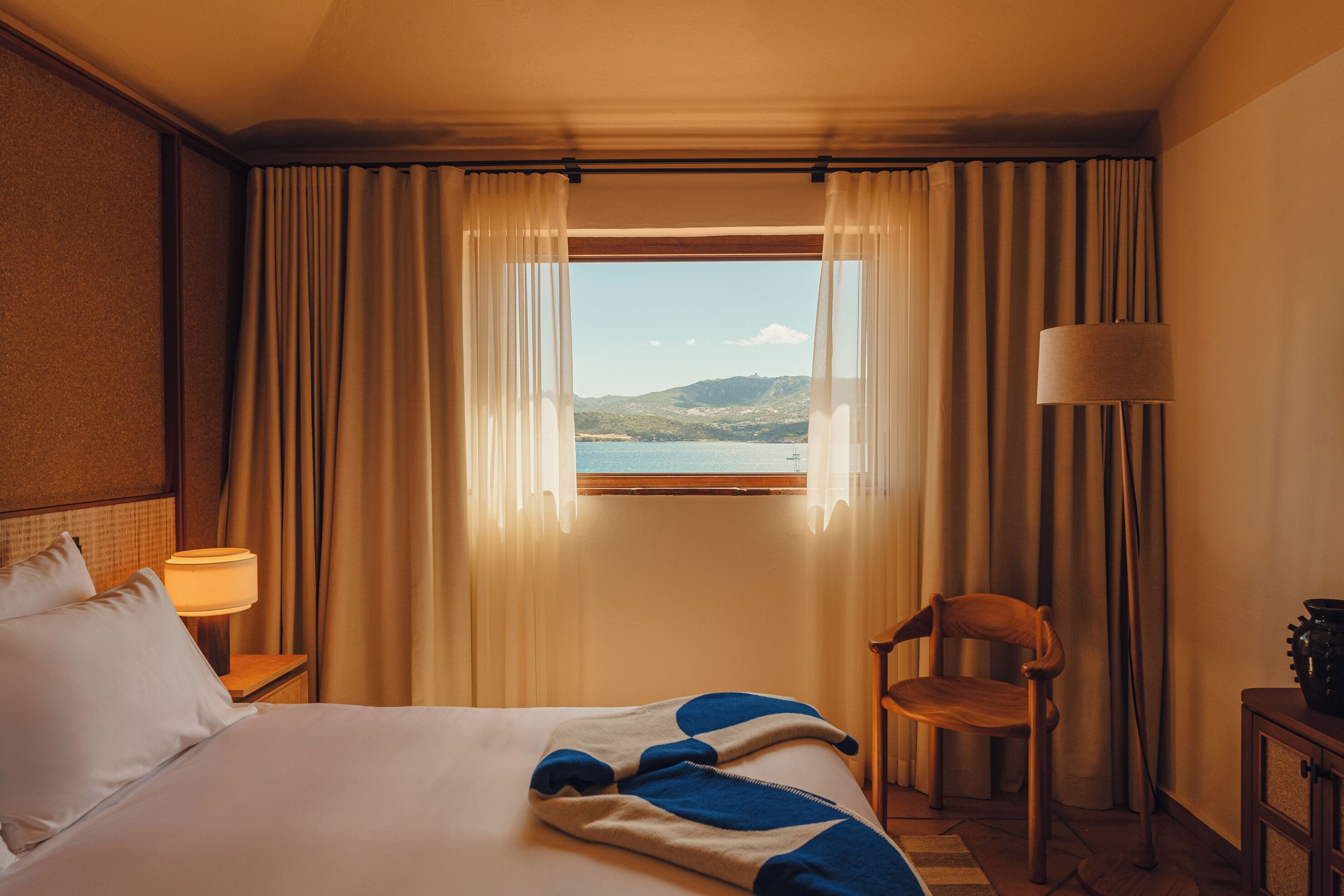 Ein Hotelzimmer mit Bett, Sessel, Lampe und Seeblick durch ein Fenster.