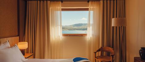 Ein Hotelzimmer mit Bett, Sessel, Lampe und Seeblick durch ein Fenster.