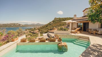 A swimming pool with clear water, surrounded by potted plants and outdoor furniture, overlooking a scenic landscape with mountains and a body of water.